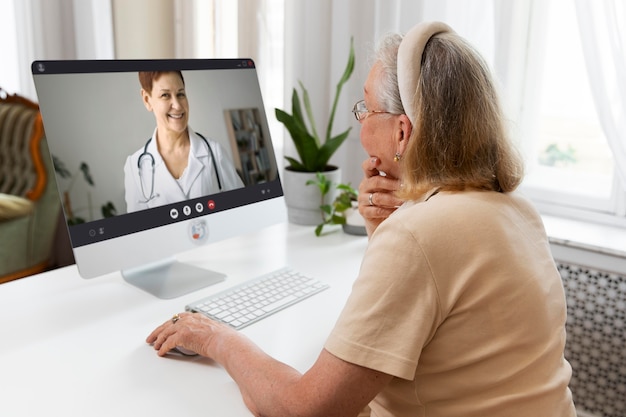A senior woman smiling happily during a telehealth appointment with her doctor. She is holding a tablet and sitting comfortably in her living room, with a smart monitor displaying her vital signs in the background. The lighting is warm and natural, and the overall scene conveys a sense of ease and accessibility.