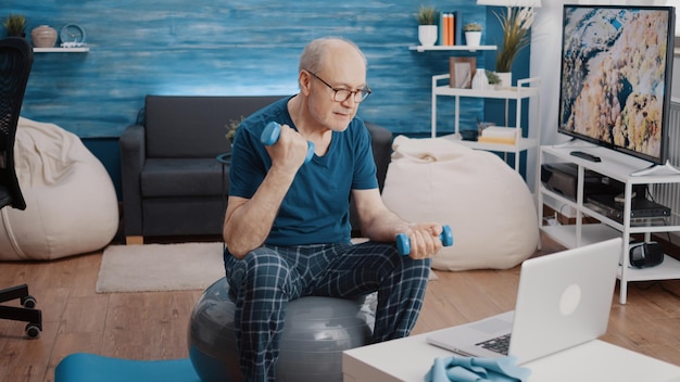 An older man in workout clothes doing squats at home with dumbbells while looking at a laptop on a table as if he is following an online workout video.