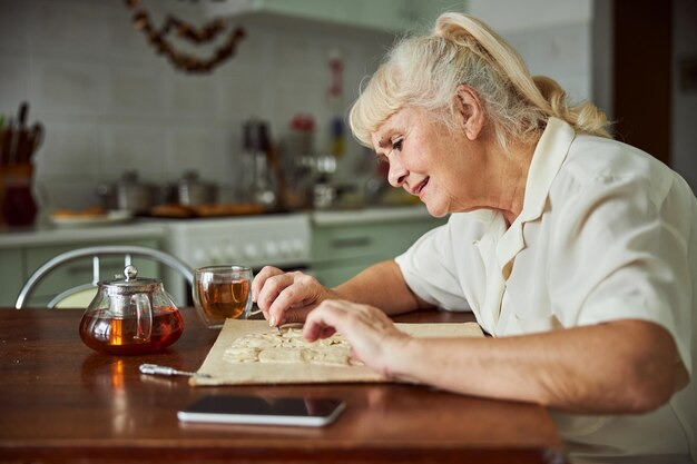 An elderly person smiling while solving a crossword puzzle at a table, with a cup of tea and a book nearby, illustrating cognitive engagement and mental stimulation.