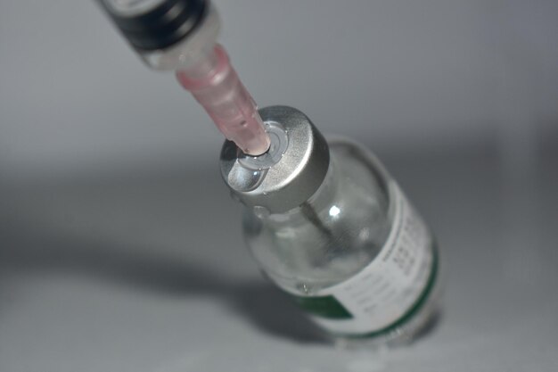 A close-up of a vial of vaccine with a needle drawing out a dose, set against a blurred background of medical supplies and equipment. The lighting is soft and professional, highlighting the precision and care involved in vaccine preparation.