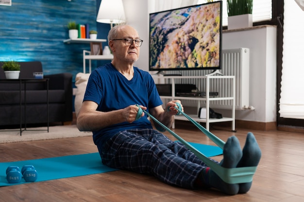 A senior man is pictured doing seated leg extensions in his living room, with a walker nearby for support. He is smiling and appears comfortable and confident.
