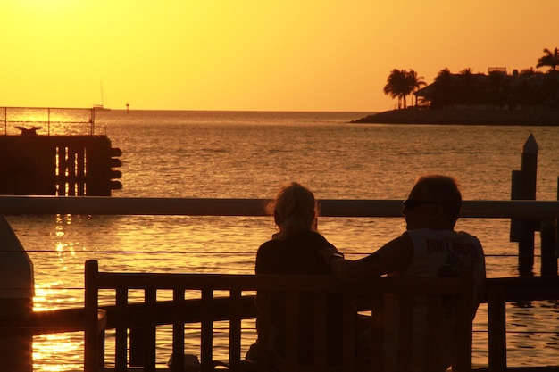 An elderly couple holding hands while walking slowly on a sandy beach at sunset. They are silhouetted against the setting sun, and the scene evokes a sense of peace, companionship, and healthy aging.