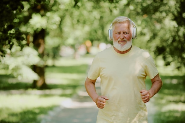 A close-up shot of a senior man smiling while exercising outdoors. He is jogging in a park with lush greenery in the background, illustrating the importance of physical activity for healthy aging.