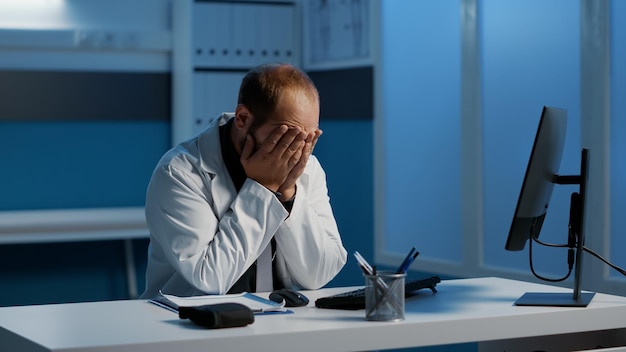 A stressed primary care physician looking overwhelmed while working late at a cluttered desk filled with paperwork and a computer screen displaying electronic health records. The image highlights the administrative burdens and long hours contributing to physician burnout.